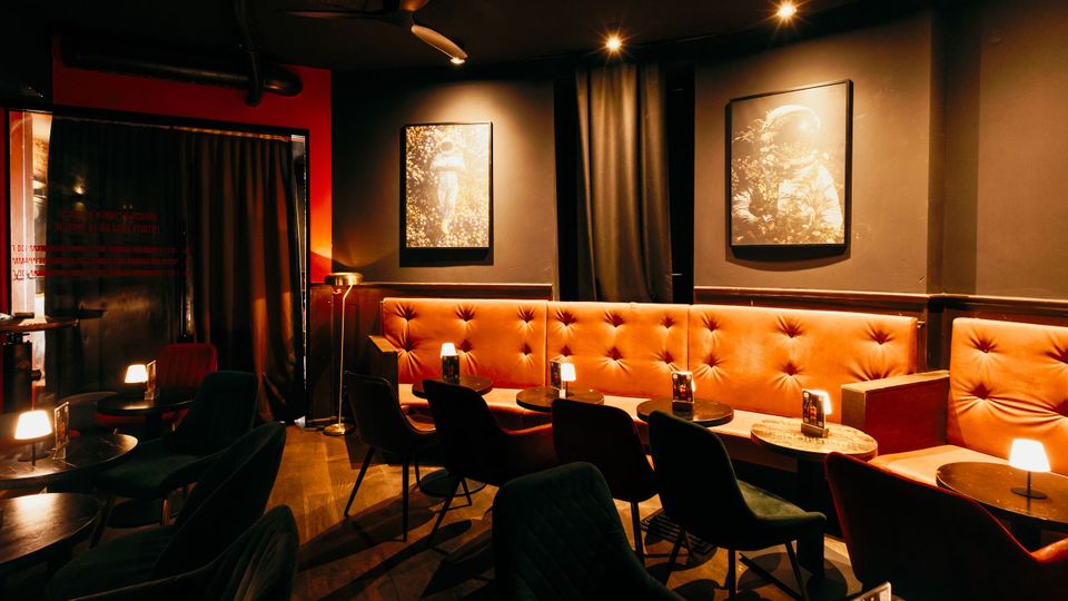Dunkel beleuchteter Bar- oder Lounge-Innenraum mit langen, orangefarbenen, gepolsterten Sitzbänken entlang der Wand, kleinen runden Tischen mit Lampen und schwarzen Stühlen. An den dunkelgrauen Wänden hängen gerahmte Bilder, und schwere Vorhänge sowie Deckenstrahler prägen die Atmosphäre.