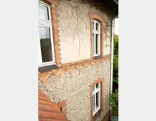 Außenansicht einer älteren Hausfassade mit hellem Putz und sichtbaren Ziegelumrahmungen um die Fenster, teils mit Bogenstürzen. An der Wand sind kletternde Ranken zu sehen, rechts verläuft ein Regenfallrohr, und unten links ragt ein Ziegeldach ins Bild.