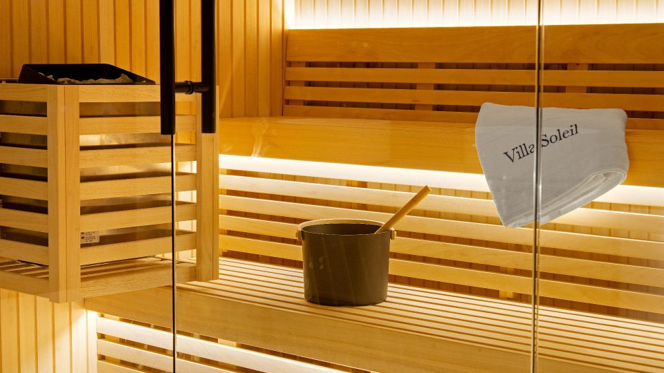 Innenansicht einer modernen Sauna mit hellen Holzlamellen, mehrstufigen Sitzbänken und warmem indirektem Licht. Links steht ein Saunaofen mit Steinen, davor ein Aufgusseimer mit Kelle; an der Glaswand hängt ein zusammengerolltes Handtuch.