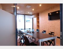 Konferenzraum mit Holzwänden und einer großen Fensterfront, die natürliches Licht hereinlässt. Ein langer Tisch mit schwarzen Stühlen, ein an der Wand montierter Fernseher und offene Regale prägen den Raum.