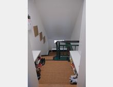 Flur mit niedriger Dachschräge, einem Treppengeländer mit grünem Metallgitter und Holzelementen. Links und rechts befinden sich weiße Schuhregale mit mehreren Paar Schuhen, der Boden ist mit einer gewebten Matte bedeckt.