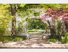 Chinesischer Garten mit weißer Wand, die von Kletterpflanzen bedeckt ist, und einem achteckigen Durchgang, der den Blick auf einen grünen Park mit Bäumen und einer Bank im Hintergrund freigibt. Im Vordergrund befinden sich Bambuspflanzen und Sonnenschatten auf dem gepflasterten Weg.