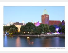Blick auf das Märkische Museum in Berlin, aufgenommen von der gegenüberliegenden Flussseite bei Dämmerung. Der rote Backsteinturm mit grünem Dach ist umgeben von Bäumen, hinter denen farbige Beleuchtungen hervorleuchten, und das ruhige Wasser des Flusses spiegelt die Lichter wider.