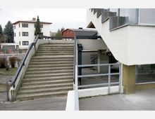 Treppenaufgang aus Beton mit Metallgeländern, der zu einer erhöhten Ebene vor einem Gebäude führt; darunter ein überdachter Bereich mit Graffiti an einer Wand. Im Hintergrund sind Wohnhäuser und Bäume sichtbar.
