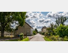 Dorfstraße mit alten Häusern und Gärten, gesäumt von Bäumen und Zäunen; im Hintergrund sind weitere Gebäude und eine von Wolken bedeckte Himmelsszenerie zu sehen.