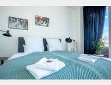 Helles Schlafzimmer mit einem Doppelbett, das mit einer türkisfarbenen Decke und weißen Kissen ausgestattet ist. An den Wänden hängen Schwarz-Weiß-Fotografien, auf einem Nachttisch steht eine schwarze Lampe, und neben dem Bett befindet sich eine Pflanze auf einem Beistelltisch.