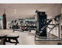 Fitnessstudio mit verschiedenen Trainingsgeräten, darunter Laufbänder, Fahrradergometer, eine Liege für Gewichtstraining und ein Boxsack. Im Hintergrund befindet sich eine Spiegelwand mit der Aufschrift "Head in the Clouds".