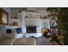 Wohnraum mit einem zentralen, weißen Kachelofen mit integrierter Sitzbank und Holzlager. Im Vordergrund befindet sich ein beiges Ledersofa, während im Hintergrund ein Mann in einem angrenzenden Raum zu sehen ist; Pflanzen und Dekorationsgegenstände ergänzen das Bild.