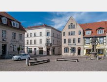 Historischer Marktplatz mit gepflastertem Boden, umgeben von mehrstöckigen Gebäuden mit schrägen Dächern und klassischen Fassaden. In der Mitte steht eine Statue auf einem Brunnen, daneben befinden sich Sitzbänke und ein geparktes Auto.