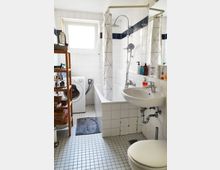 Badezimmer mit weiß gefliesten Wänden und grauem Fliesenboden, ausgestattet mit einer Badewanne mit Duschvorhang, einer Waschmaschine, einem weißen Waschbecken und einem WC. Ein Holzbordregal steht an der linken Wand, und ein Fenster sorgt für Tageslicht.