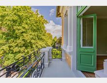 Schmaler Balkon an der Fassade eines Wohnhauses mit weißer Stuckverkleidung, Ziegelwand und schwarzem schmiedeeisernem Geländer. Eine offene grüne Balkontür führt in den Innenraum; neben dem Balkon stehen hohe, dicht belaubte Bäume.