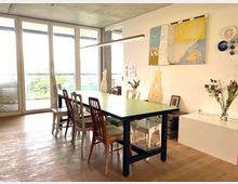 Esszimmer mit großem rechteckigem Tisch und verschiedenen Stühlen, modernem Holzboden und einer Wand mit Kunstwerken und dekorativen Elementen. Große Fensterfront mit Blick ins Freie lässt viel Tageslicht herein.