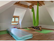 Dachgeschossraum mit Holzboden und sichtbaren Holzbalken. Im Raum befinden sich eine aufblasbare Gymnastikmatte, eine grüne Hängematte zur Luftakrobatik, sowie Fitnessgeräte auf grünen Matten vor einem Fenster mit Blick nach draußen.