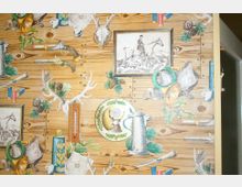 Holzvertäfelte Wand mit jagdlichem Dekor, darunter Illustrationen von Hirschgeweihen, Jagdhörnern, Pistolen und Bildern von Jägern. Integriert sind ein Thermometer und Dekorelemente mit Naturmotiven wie Tannenzweigen und Kiefernzapfen.