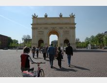 Eine historische Triumphbogenstruktur auf einem großen gepflasterten Platz, mit Personen, die zu Fuß oder mit dem Fahrrad in Richtung des Bogens unterwegs sind. Im Hintergrund befinden sich Bäume und Gebäude, die den Platz umgeben.