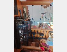 Innenraum eines gemütlich eingerichteten Wohn- oder Aufenthaltsbereichs mit Holzverkleidung, einer Bar mit hohen schwarzen Hockern und Pendelleuchten. Im Vordergrund ist ein Schrank mit Glastüren zu sehen, der mit Dekoobjekten und Büchern gefüllt ist.