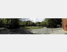 Panorama eines kleinen Stadtparks mit einem zentralen Blumenbeet in Kreisform, umgeben von gepflasterten Wegen und Bäumen. Im Hintergrund sind Wohngebäude zu sehen, rechts im Bild ist teilweise eine rote Backsteinfassade eines Gebäudes erkennbar.