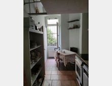 Kleine Küchenzeile mit offenen Regalen, gefüllt mit Geschirr. Im Hintergrund steht ein Esstisch mit gehäkelter Tischdecke und zwei Stühlen, daneben ein Fenster mit Gitter und Blick ins Grüne.