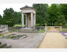 Kleiner klassizistischer Pavillon mit Säulen und einer Büste im Inneren, umgeben von Steinplatten, Zierbeeten und einem gepflegten Garten mit Bäumen und einem schmiedeeisernen Tor. Die Umgebung ist geprägt von grünem Rasen und hohen Bäumen.