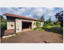 Ein flaches Gebäude mit beiger Fassade und rotem Satteldach, dessen offene Garage Werkzeuge und Gegenstände enthält. Der Vorderhof besteht aus gepflasterten Steinen, umgeben von Rasenflächen, Bäumen und einem Zauntor im Hintergrund.