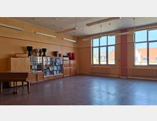 Großer, leerer Musik- oder Proberaum mit Linoleumboden, hohen Deckenleuchten und großen Fenstern mit Heizkörpern darunter. An der Wand stehen Holzschränke und Vitrinen mit Instrumenten, daneben ein Klavier; durch die Fenster sind Dächer benachbarter Gebäude zu sehen.