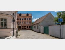 Eine gepflasterte Gasse mit historischen Gebäuden, darunter ein rosafarbenes Mehrfamilienhaus mit dekorativen Fassadenelementen und zwei kleinere Häuser mit roten Ziegeldächern und grün gestrichenen Türen und Fensterläden. Ein Straßenschild weist auf die „Altstädtischer Kietz“-Zone hin.