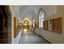 Langer, heller Korridor mit gewölbter Decke, seitlichen Schaukästen aus Holz und hohen Fenstern auf der rechten Seite. Links im Bild sind Teile einer braunen Steinumrahmung sichtbar.