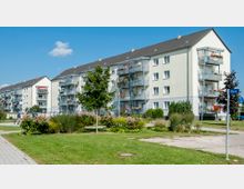 Mehrstöckige Wohngebäude mit hellen Fassaden und Balkonen, umgeben von gepflegten Grünflächen mit Bäumen, Sträuchern und Blumen. Im Vordergrund verlaufen Gehwege, und ein Straßenschild mit der Aufschrift "Uferstr." ist sichtbar.