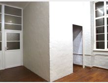 Ein heller Innenraum mit weißen Wänden, einem Holzboden und großen Fenstern mit Sprosseneinteilung. Rechts ist ein Durchgang zu einem weiteren Raum sichtbar, links eine weiße Tür mit Milchglaseinsätzen.