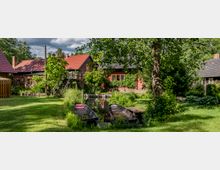 Ländliche Szene mit mehreren Häusern in traditioneller Bauweise, umgeben von üppigem Grün und Bäumen. Im Vordergrund liegt ein kleiner Wasserkanal mit zwei Holzbooten, hinter dem sich eine gepflegte Wiese erstreckt.