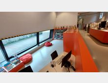 Innenraum einer modernen Bibliothek mit orangefarbenen Möbeln, darunter ein gebogener Tresen und Bücherregale. Große Fenster mit Blick auf einen Gehweg lassen Tageslicht herein, neben ihnen liegen rote Sitzsäcke auf hellem Boden.