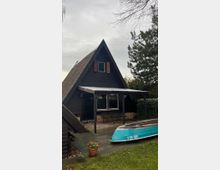 Ein kleines, dunkelbraunes A-Frame-Haus mit Spitzdach, einer überdachten Terrasse und einem großen Fenster mit Fensterläden. Im Vordergrund steht ein blaues Ruderboot auf einem gepflasterten Bereich, daneben ein Blumentopf mit gelben Blumen, umgeben von Rasen und Bäumen.