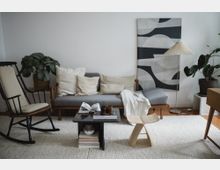 Ein gemütliches Wohnzimmer mit einem grauen Sofa, hellen Kissen und einer Decke, daneben eine Stehlampe und große Zimmerpflanzen. Im Vordergrund steht ein niedriger Couchtisch mit Büchern und Gläsern, ein Holzhocker und ein schwarzer Schaukelstuhl auf einem hellen Teppich.