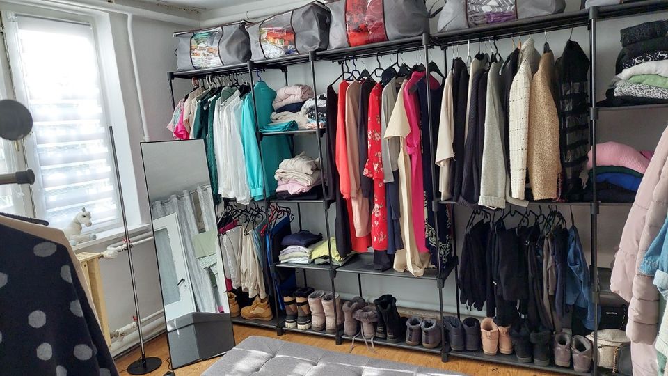 Offenes Ankleidezimmer mit freistehendem Metall-Kleiderregal, an dem zahlreiche Kleidungsstücke auf Bügeln hängen, sowie Ablagefächern mit gefalteter Wäsche, Schuhreihen und Aufbewahrungsboxen. Daneben stehen ein großer Standspiegel und ein Fenster mit Jalousie in einem hellen Raum mit Holzboden.