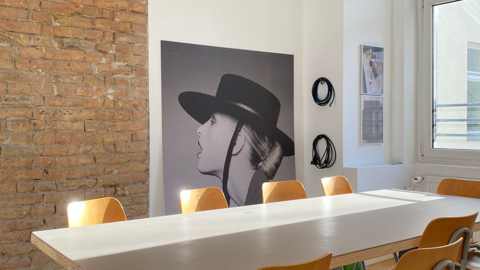Heller Besprechungsraum mit langem rechteckigem Tisch und mehreren Holzstühlen auf Teppichboden, links eine freiliegende Backsteinwand. An der weißen Wand lehnt ein großes Schwarzweiß-Porträt, darüber hängt eine runde Papier-Pendelleuchte; rechts sorgt ein großes Fenster für viel Tageslicht.