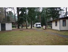 Eine Waldlandschaft mit mehreren kleinen, einfachen Gebäuden mit verputzten Wänden. In der Mitte steht ein weißes Wohnmobil, umgeben von hohen Nadelbäumen und vereinzelten Gehwegen.