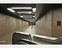 Unterirdischer Fußgängertunnel mit roten Ziegelwänden, gefliestem Boden und länglichen Deckenleuchten. Im Hintergrund sind mehrere Personen zu Fuß unterwegs, während im Vordergrund ein Teil einer Rolltreppe sichtbar ist.