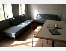 Minimalistisch eingerichteter Raum mit grauen, gepolsterten Sofas in L-Form an einer Wand mit großen Fenstern. Vor den Sofas steht ein Tisch aus Holz mit Metallbeinen, darauf liegen zwei offene Bücher; ein Holzstuhl steht daneben.