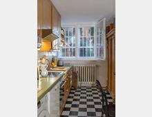 Eine kleine, helle Küche mit Holzmöbeln, schwarz-weiß kariertem Fliesenboden und einer großen, zweiflügeligen Fensterfront. Rechts steht ein Heizkörper unter den Fenstern, links befinden sich ein Herd, eine Spüle und Arbeitsflächen mit Geräten wie einem Wasserkocher und einer Kaffeemaschine.