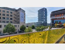 Städtische Ansicht mit modernen Gebäuden, darunter ein Bürogebäude mit hellem Stein und ein Hochhaus im Hintergrund. Im Vordergrund eine gelbe Brüstung mit aufgedruckten Smileys; rechts ein Einkaufszentrum mit dem Schriftzug „EAST“ und Außenbereich mit gelben Sonnenschirmen.