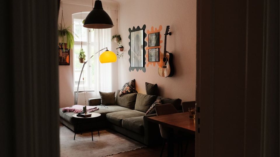 Gemütliches Wohnzimmer mit großer Eckcouch, kleinem runden Couchtisch und Stehlampe neben einem hohen Fenster mit hängenden Pflanzen. An der Wand hängen dekorative Spiegel und eine Gitarre, dazu eine große Pendelleuchte über dem Sitzbereich.