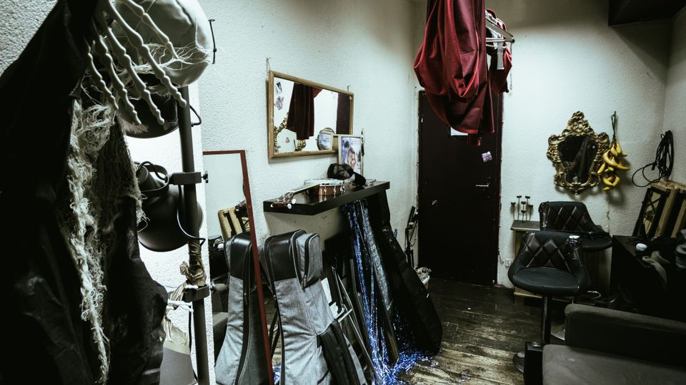 Small, cluttered backstage dressing room with white textured walls, wooden floorboards, and a vanity shelf beneath a wall mirror. Costumes and props hang from hooks and a ceiling rack, with instrument cases and frames stacked along the walls near a black door and ornate mirrors.