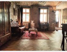 Ein Wohnzimmer mit einer Ziegelwand, großen Fenstern und einem hölzernen Fischgrätenboden. Das Interieur umfasst ein Ledersofa, einen Holztisch mit einer Vase, einen roten Teppich und antike Möbel, darunter einen Schrank und Stühle.