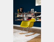 Gelber Schaukelstuhl mit Metall- und Holzgestell auf einem weißen Teppich vor einer dunkelblauen Wand. Im Hintergrund ein Regal mit Büchern, Brettspielen und Dekorationsgegenständen.