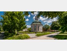 Barocke Kapelle mit pastellfarbenem Putz und einer markanten grünen Kuppel, umgeben von einem parkähnlichen Garten mit gepflasterten Wegen, Sitzbänken und schattenspendenden Bäumen, unter einem blauen Himmel mit vereinzelt weißen Wolken.