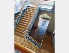 Holztreppe mit verzierten, hellblauen Geländern in einem Innenraum mit Parkettboden und einer offenen Galerie. Im Hintergrund ist ein Durchgang mit Fliesenboden und einer weißen Tür erkennbar.