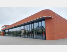 Modernes Gebäude mit einer Fassade aus roten Klinkersteinen und großen, vertikal ausgerichteten Glasfenstern, die eine reflektierende Oberfläche haben; das Gebäude hat abgerundete Ecken und steht auf einem gepflasterten Platz. Im Hintergrund sind Bäume und Wohnhäuser zu sehen.