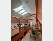 Dachgeschosswohnung mit sichtbaren Holzbalken, großen Dachfenstern, die Tageslicht hineinlassen, und einem offenen Wohnbereich mit roten Fliesen, einer Küche mit Holzarbeitsplatte, einer Sitzecke mit Sofa und einem Esstisch im Vordergrund. Im Hintergrund führen Türen zu weiteren Räumen.