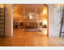 Ein rustikaler Wohnraum mit Holzboden, einer schlichten Tapete mit abgenutzter Optik, einer antiken Couch und zwei Stühlen, einem zentral platzierten Teppich und dezenter Beleuchtung durch Wandlampen. Links steht eine Holzleiter, und ein Spiegel hängt über der Couch.