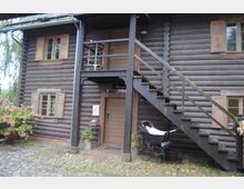 Holzhaus mit Blockbohlenfassade, einem Eingang im Erdgeschoss und einer Holztreppe, die zu einem oberen Balkon mit Tür führt. Im Vordergrund ein gepflasterter Hof, ein alter Kinderwagen und blühende Pflanzen in Töpfen.